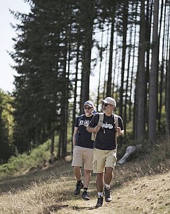 Two men are hiking on a path in Bourscheid during the summer. In the background, there is a fir forest. The men are dressed in summer attire, wearing shorts and black shirts.