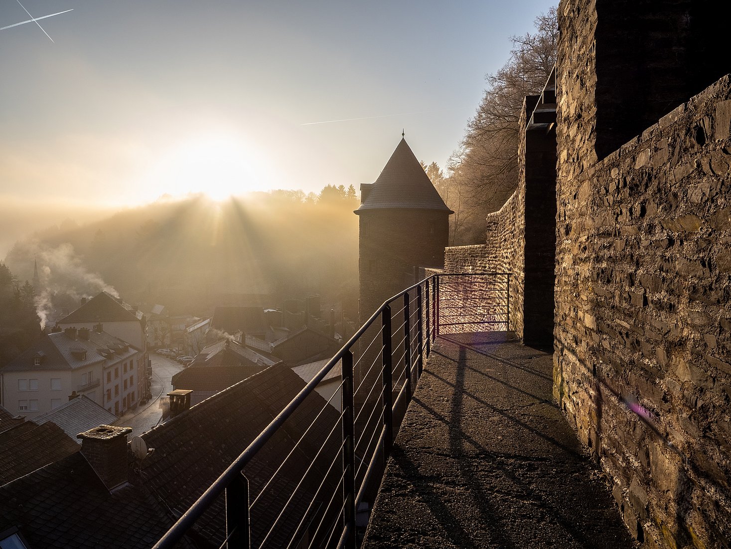 A photo taken at a fortress wall, with the sunrise casting light over the castle and highlighting the small tower against the blue sky.