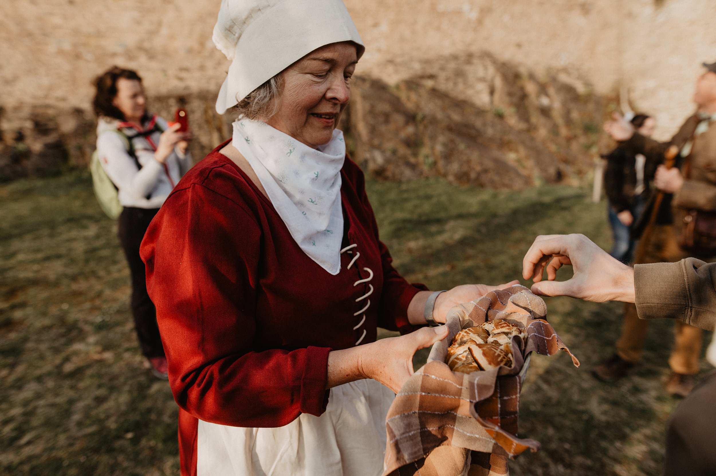 Une femme vêtue d'une robe rouge et d'un tablier blanc sert de la nourriture à un groupe de visiteurs devant un château.