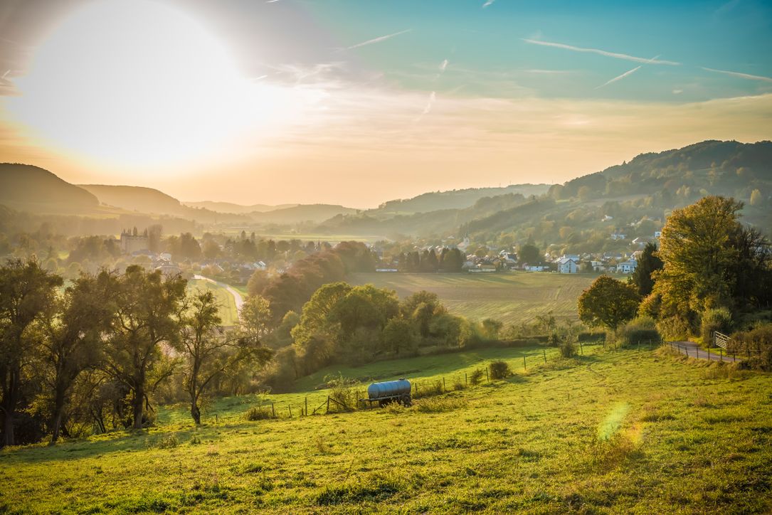 Un paysage vallonné au printemps, entouré de forêts et de champs, où la ville dans la vallée se mélange à une légère brume, tandis que le ciel est bleu.