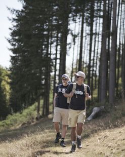 Twee mannen wandelen in de zomer over een pad in Bourscheid. Op de achtergrond is een dennenbos te zien. De mannen zijn zomers gekleed in shorts en zwarte shirts.