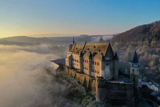 Een droneshot van kasteel Vianden, omringd door mist. Ondanks de mist is de zon zichtbaar en de blauwe lucht te zien.