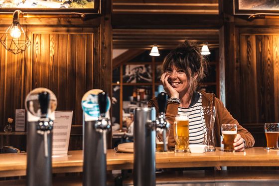 Une femme est assise au comptoir du restaurant Cornelyshaff, sourit détendue à la caméra et a une bière devant elle.