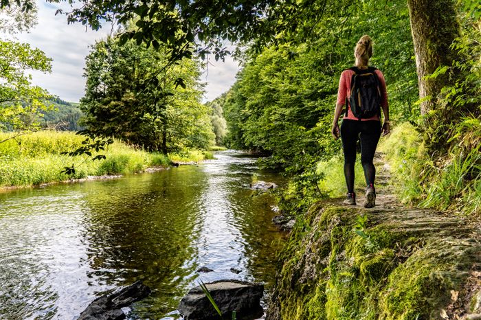 A female hiker walks along the river on the Éislek Pad Kalborn, surrounded by peaceful nature.