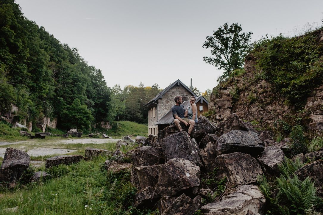 A couple sits on a rock formation, with a house in the background. The area is a nature reserve, surrounded by forest and rocks.