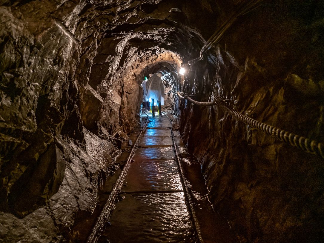 A photo in the copper mine of Stolzemburg, showing a narrow, slightly damp tunnel. In the background, lights are visible in the tunnel, which appears dark and cramped.