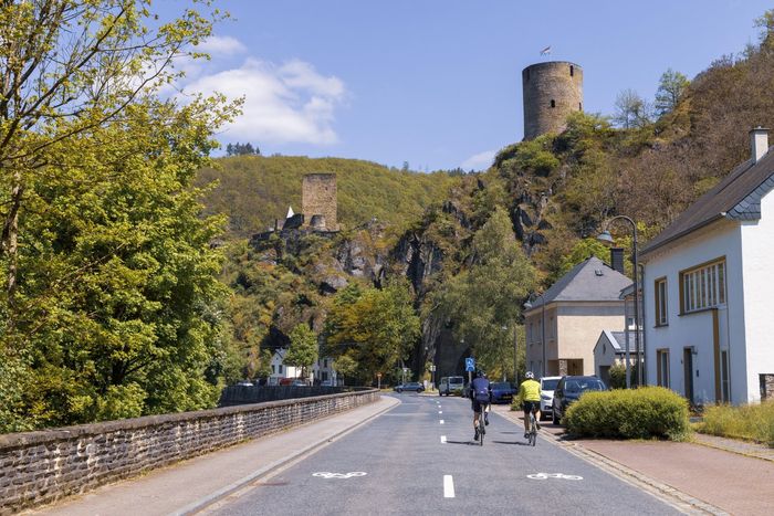 Deux cyclistes roulent côte à côte sur une route à Esch-sur-Sûre. Une partie du château d’Esch-sur-Sûre est visible en arrière-plan.