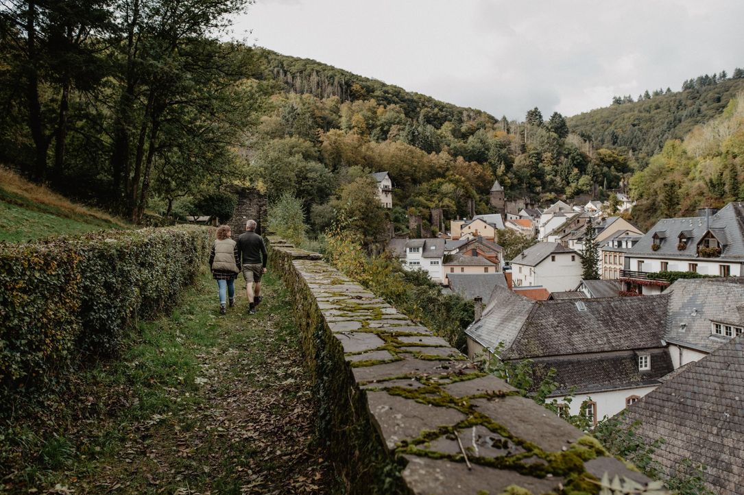 A couple walking along a moss-covered stone wall in a historic town, surrounded by green hills and traditional houses.