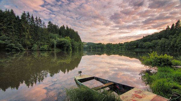 Un petit bateau est garé sur la rive de la Sûre, entouré d'une forêt dense, alors que le soleil se couche lentement.