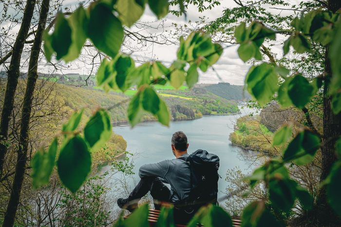 Wanderer mit Rucksack blickt durch Bäume und Vegetation auf den Obersauer-Stausee