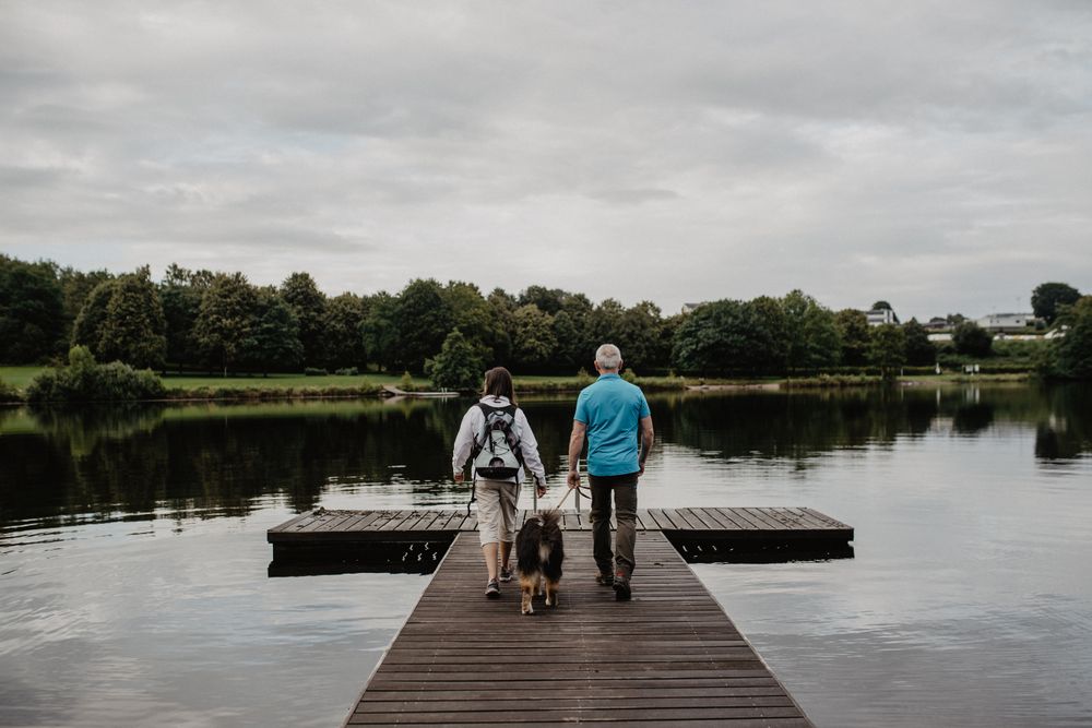 An elderly couple with a dog strolls along a wooden walkway toward the lake in Weiswampach.
