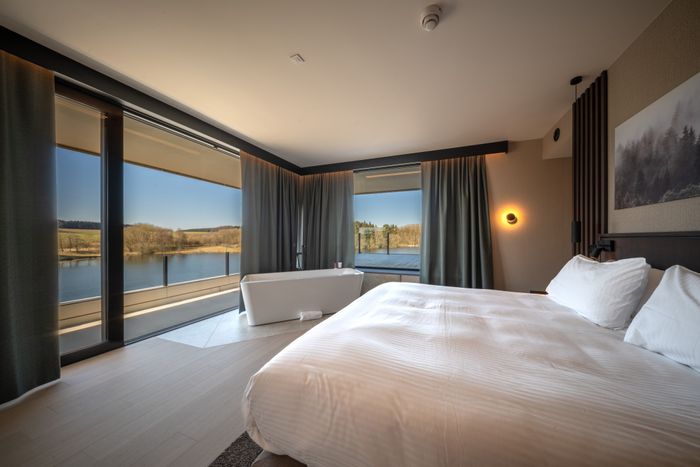 Une chambre d’hôtel à l’Hôtel Anatura à Weiswampach avec vue sur le lac de Weiswampach à travers de grandes fenêtres.