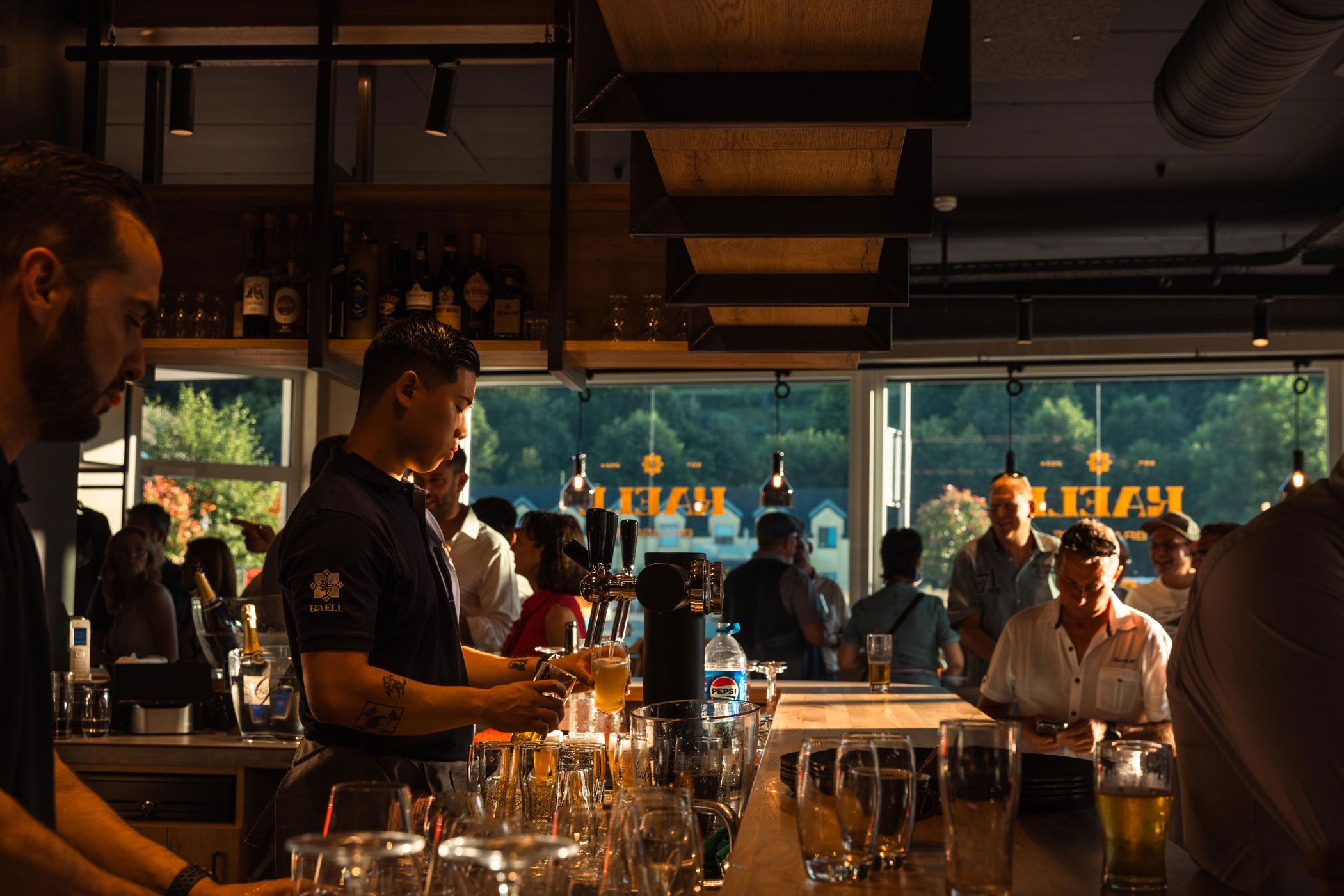 A bartender preparing a drink stands in front of the panoramic window at the Kaell Bistrot-Brasserie in Ettelbruck, with the sunset visible in the background.