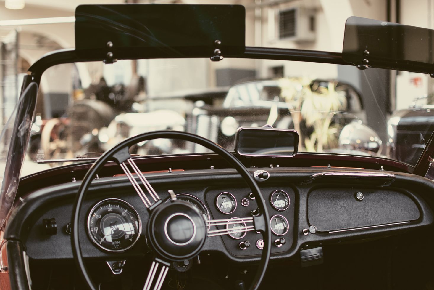 Een binnenaanzicht vanuit een auto waarin het stuur van een oude cabriolet te zien is. Andere vintage auto's zijn te zien door het raam, maar ze zijn niet duidelijk zichtbaar.