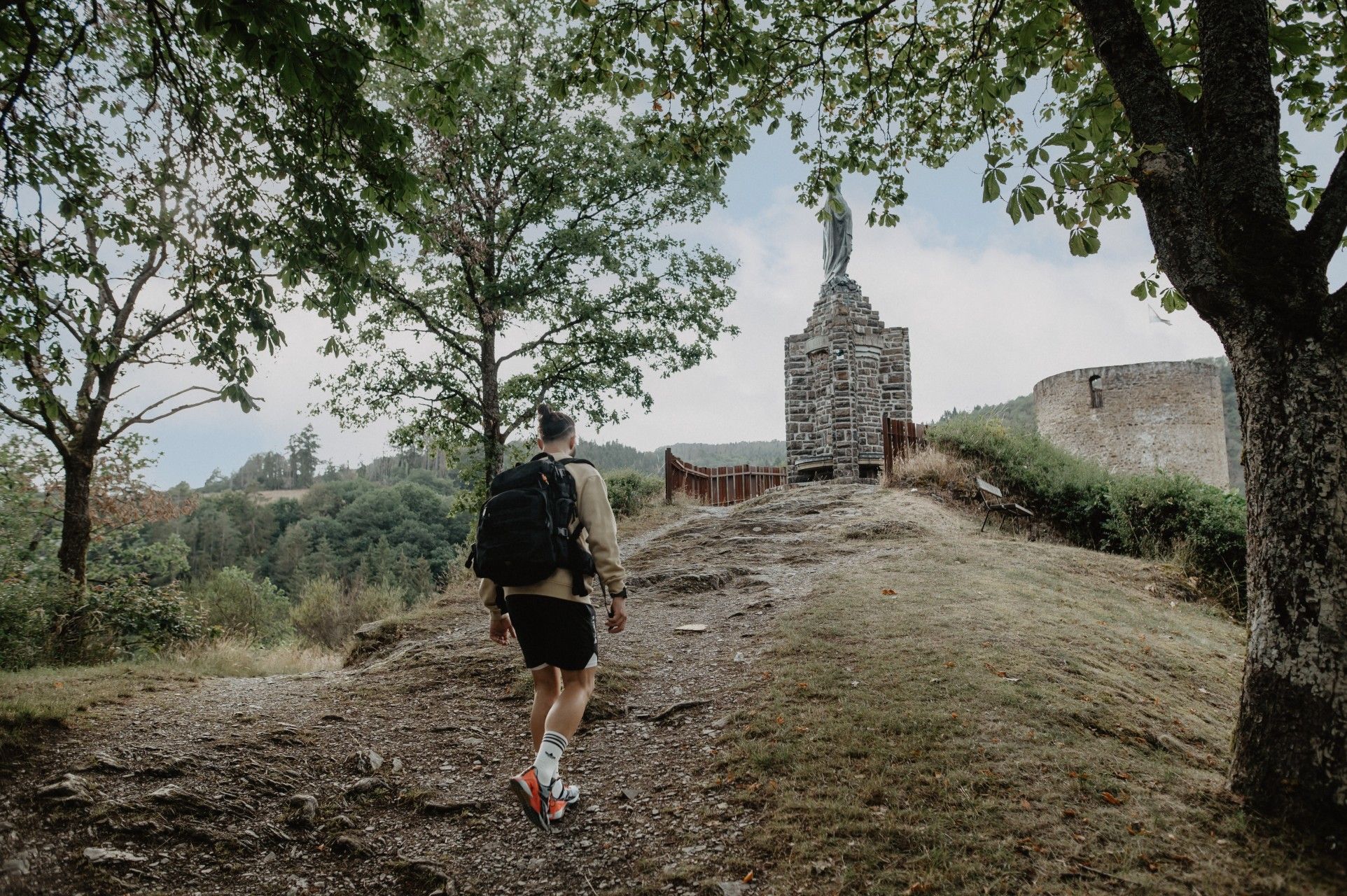 Hiker walking uphill on the Éislek Pad near Esch-sur-Sûre with chapel and trees along the path