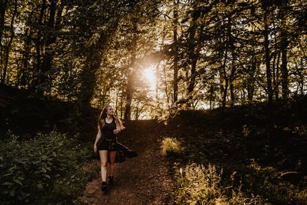 Een vrouw met blond haar, gekleed in zwart, loopt een bospad naar beneden op het Éislek Pad in Gilsdorf. De zon gaat onder en schijnt door de bladeren.