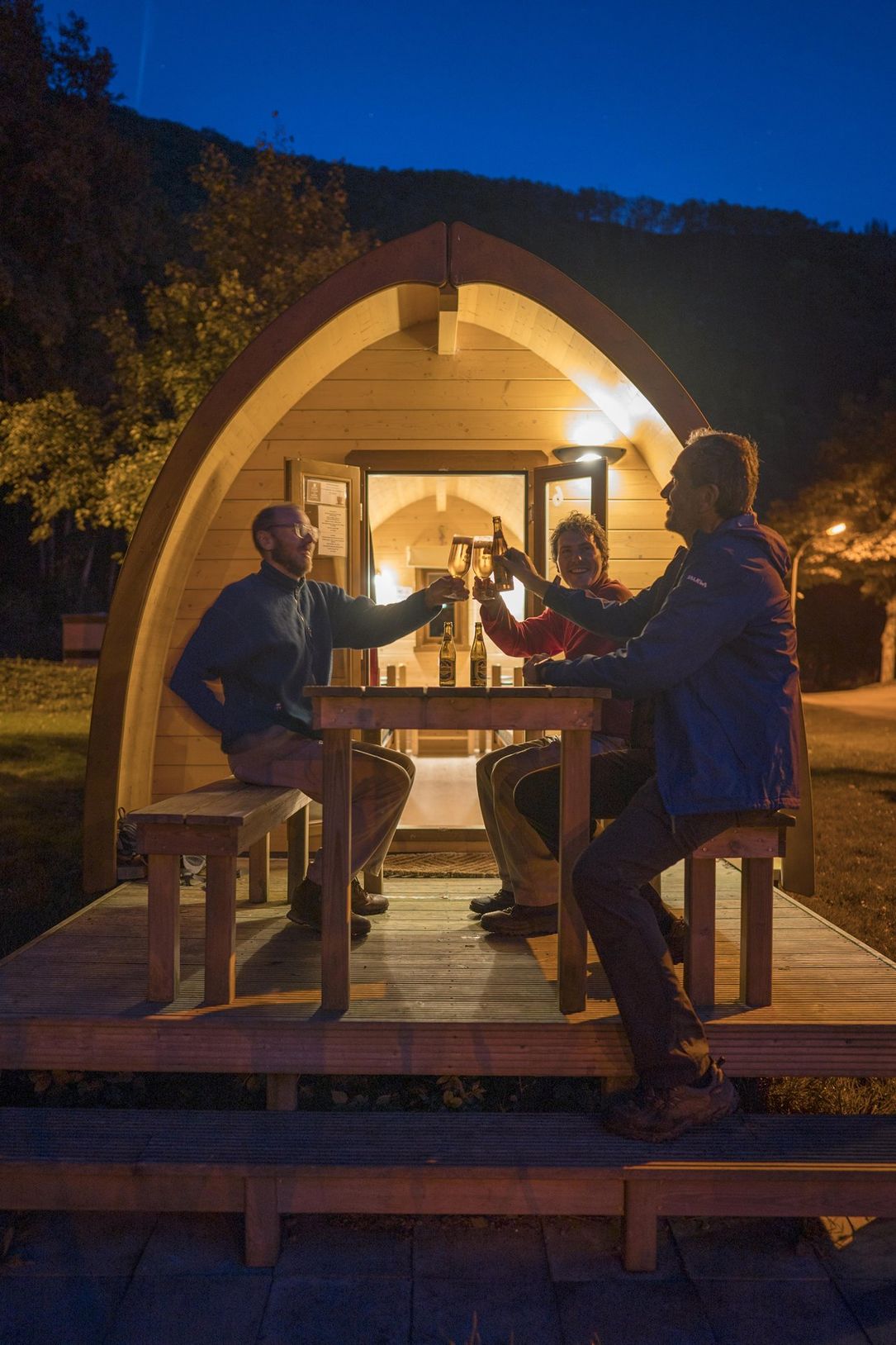 Men relaxing with a drink in front of a wooden pod at a campsite in Éislek, Luxembourg, after a day of hiking. 
