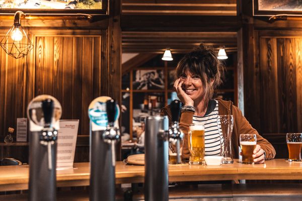Een vrouw zit aan de bar van restaurant Cornelyshaff, glimlacht ontspannen naar de camera en heeft een bier voor zich staan.