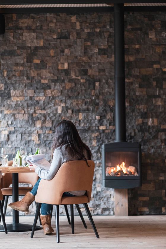 Een vrouw zit in een restaurant en leest de menukaart in een gezellige sfeer, met een vuur dat brandt in de open haard op de achtergrond.