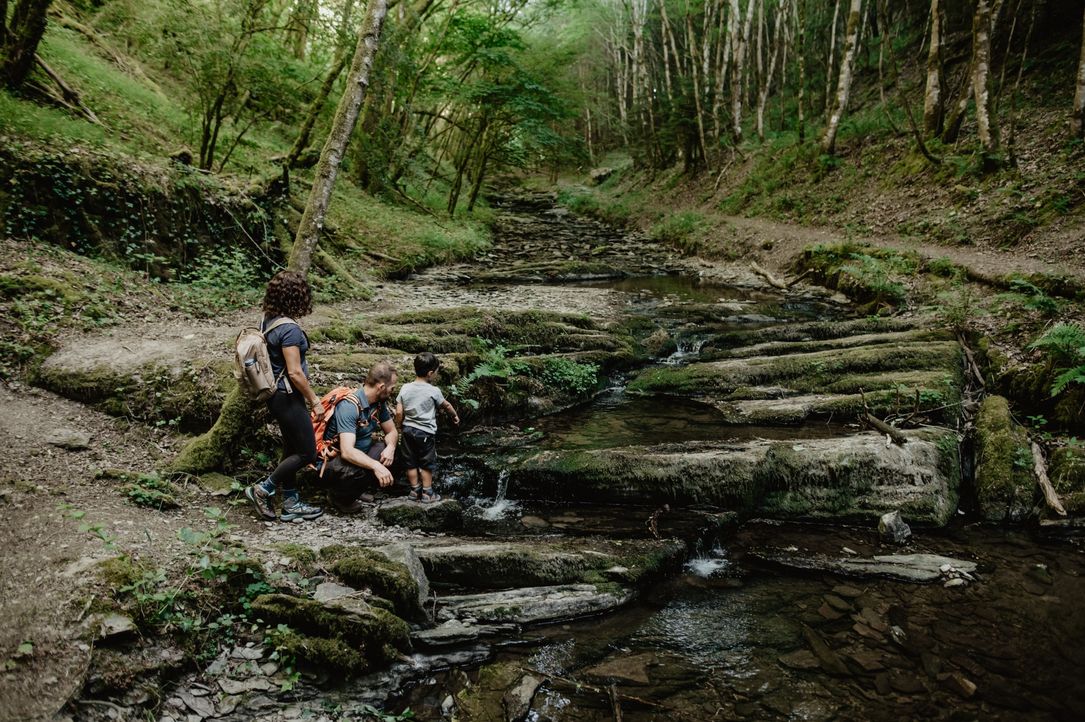Alt-Text Deutsch: Eine Familie mit einem kleinen Kind spielt an einem kleinen Wasserfall entlang des Wassersënneswee in Eschdorf.