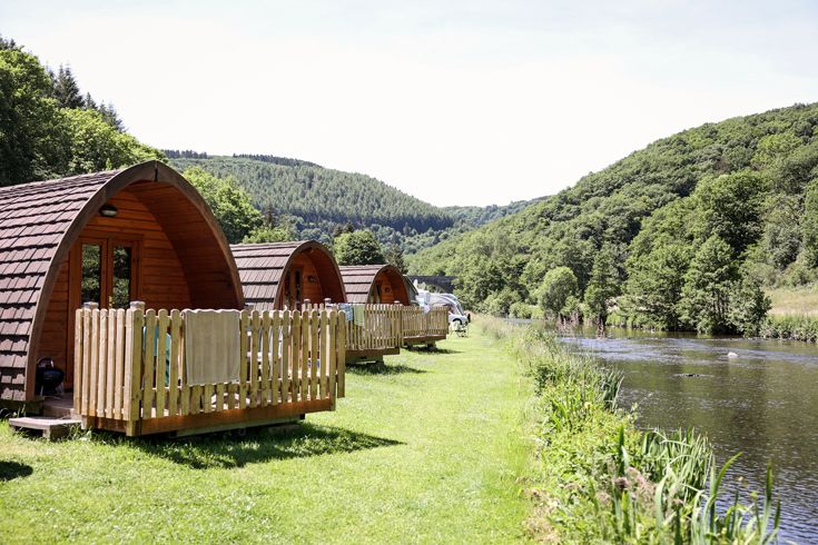 Ein Foto von drei kleinen Holzhütten des Campingplatzes Bissen, der sich im Heiderscheider Grund im Grünen und von Wald umgeben befindet, bei Sonnenschein.