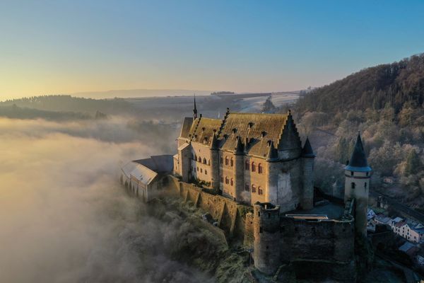 A drone shot of Vianden Castle, surrounded by mist. Despite the fog, the sun is visible and the blue sky can be seen.