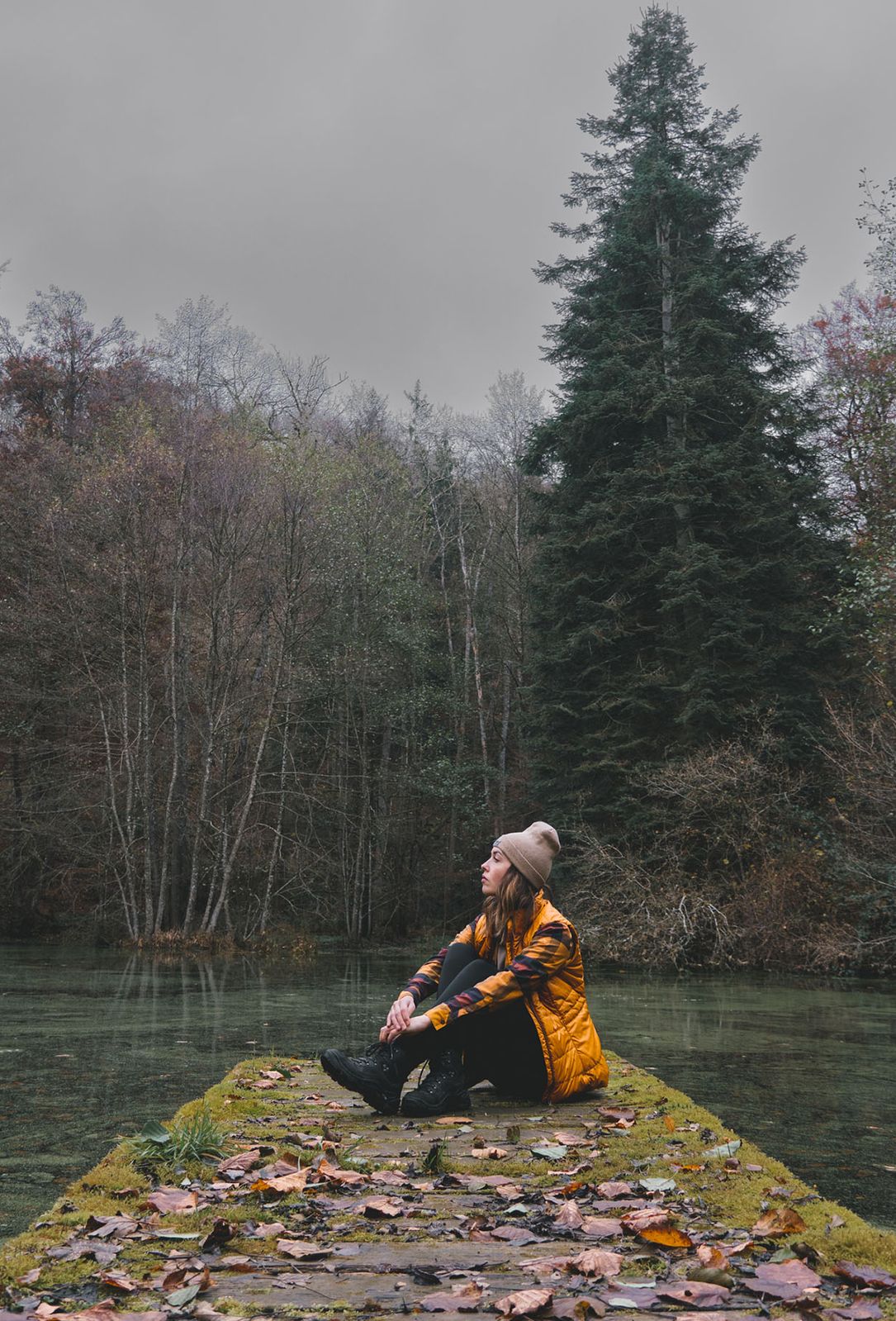 A woman sits in autumn clothing on a dock by the water, surrounded by trees that have already lost some leaves. The sky is gray.