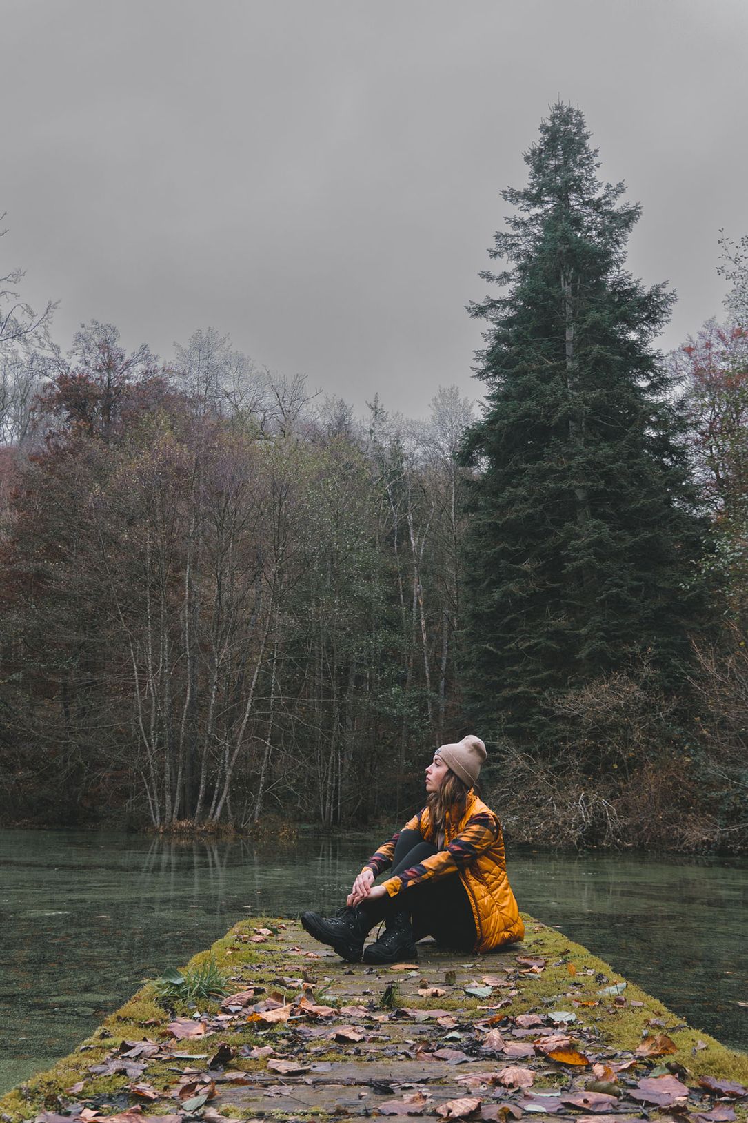 Une femme est assise en vêtements d'automne sur un ponton au bord de l'eau, entourée d'arbres qui ont déjà perdu quelques feuilles. Le ciel est gris.