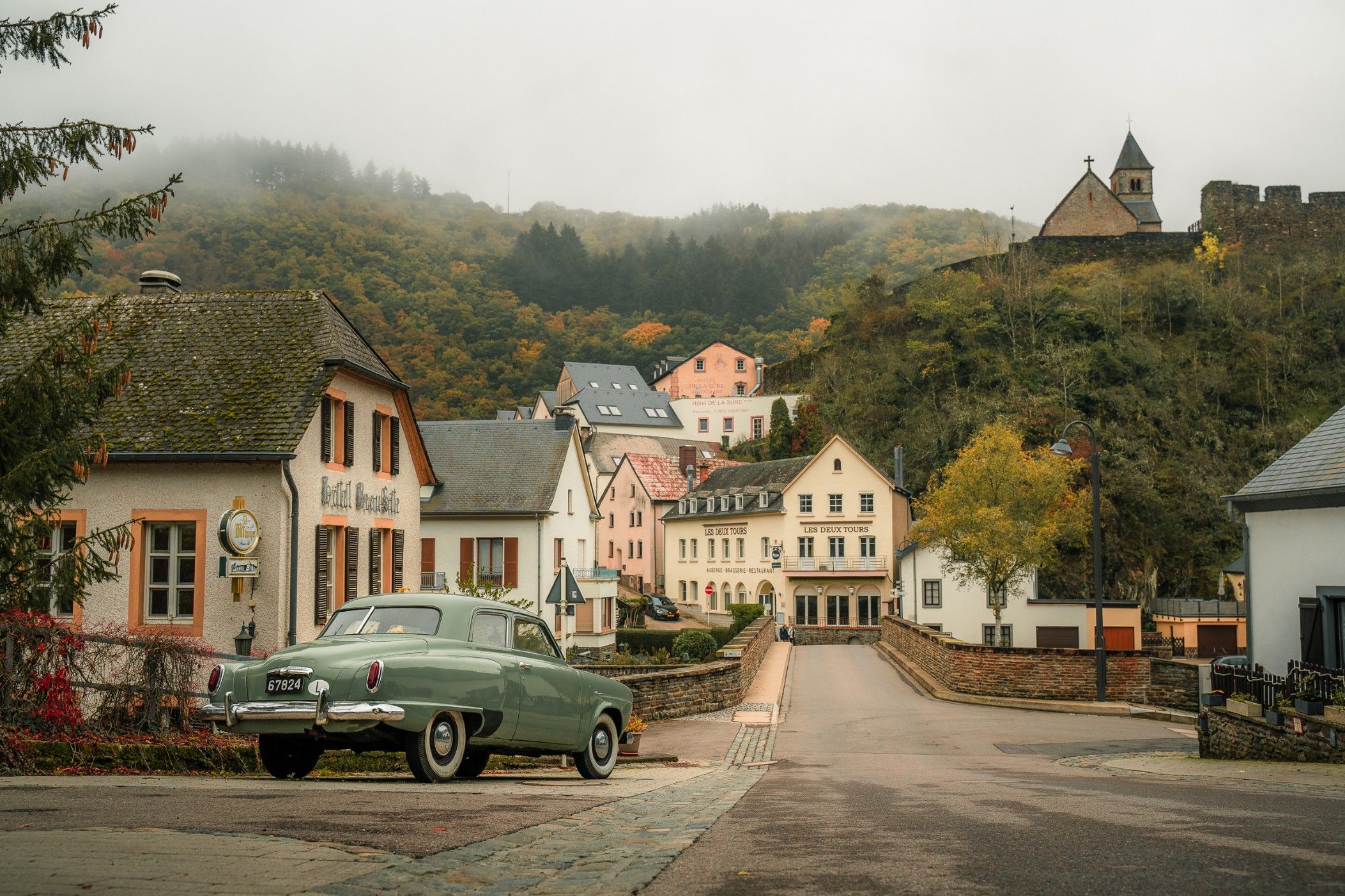 A shot of the city of Esch-sur-Sûre with a green vintage car in front of the bridge. The colorful houses and light fog provide a unique view.